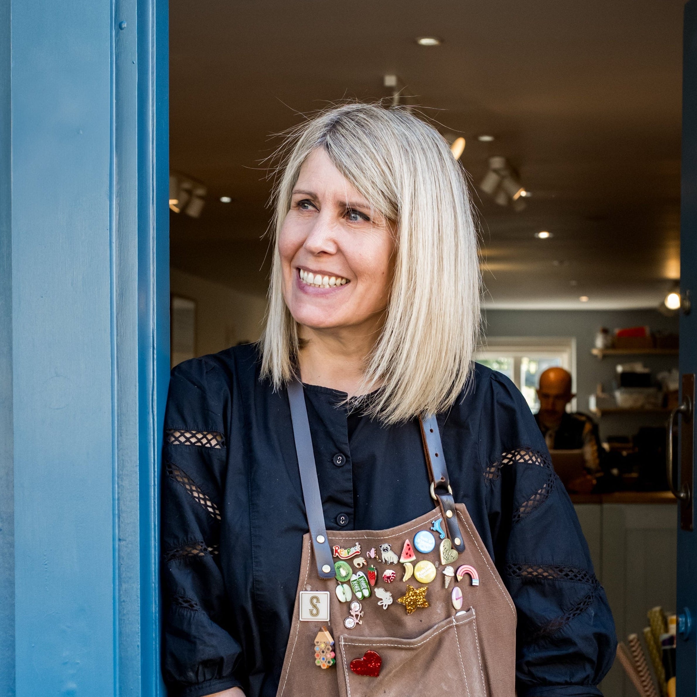 Woman wearing a brown apron with colorful buttons standing in a doorway of a store.