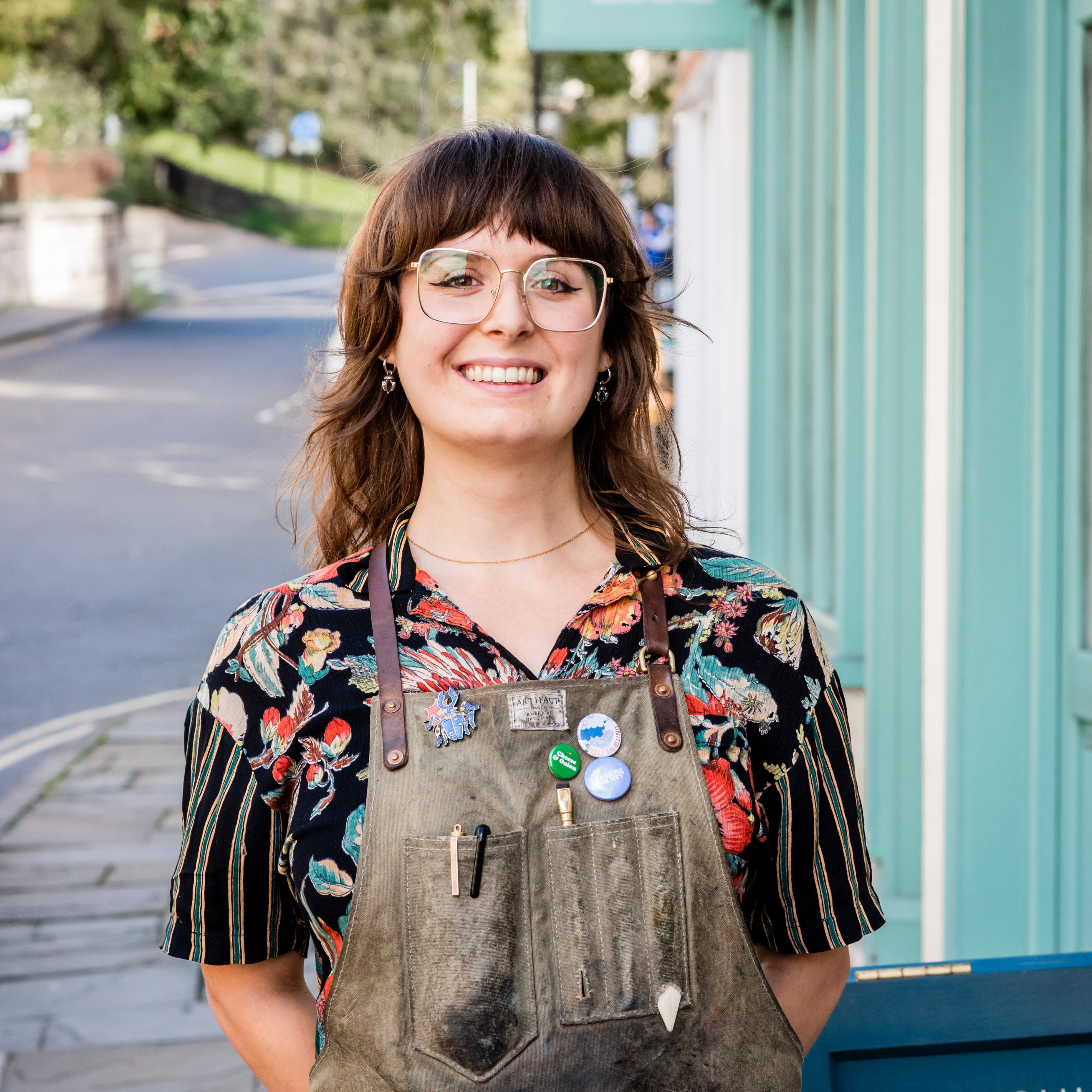 Woman wearing overalls and a floral shirt standing on a sidewalk with a building and street sign in the background.